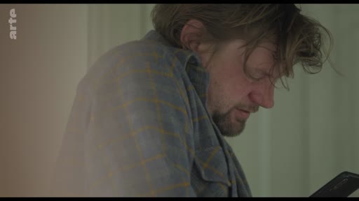 Un homme barbu, vêtu d'une chemise à carreaux, consulte son téléphone portable dans un moment d'isolement et de réflexion. L'image illustre le thème de la communication numérique et de la solitude.