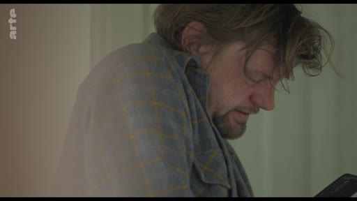 Un homme barbu, vêtu d'une chemise à carreaux, consulte son téléphone portable dans un moment d'isolement et de réflexion. L'image illustre le thème de la communication numérique et de la solitude.