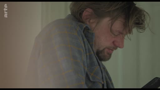 Un homme d'âge moyen, portant une chemise à carreaux, consulte son téléphone portable avec un air concentré ou soucieux. L'image illustre le thème de la communication isolée et de l'usage des technologies numériques au quotidien.