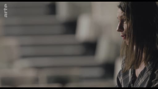 Une femme est filmée en gros plan lors d'une discussion sur un collègue, dans un cadre intérieur sobre.