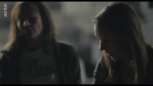 Deux jeunes femmes sont filmées en plan moyen lors d'une conversation intime dans un environnement sombre, illustrant une scène de dialogue.