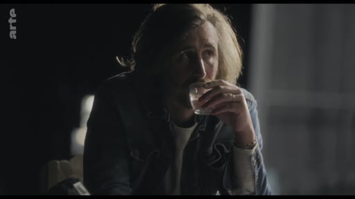 Un homme aux cheveux longs, vêtu d'une veste en jean, est filmé en gros plan alors qu'il boit dans un verre au cours d'une discussion.