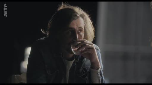 Un homme aux cheveux longs, vêtu d'une veste en jean, boit un verre tout en participant à une conversation dans une ambiance tamisée.