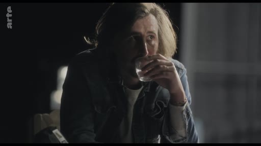 Un homme aux cheveux longs, vêtu d'une veste en jean, boit un verre dans une ambiance tamisée lors d'une séquence de portrait ou d'interview.