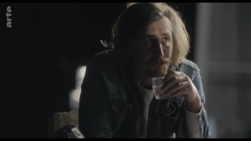 Un homme aux cheveux longs et à la barbe, vêtu d'une veste en jean, est assis dans une pièce sombre et tient un verre à la main lors d'une conversation.