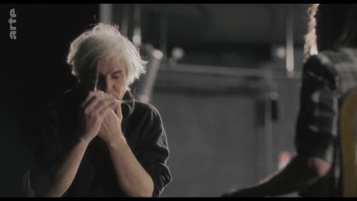 Un homme aux cheveux blancs, possiblement un acteur ou un metteur en scène, est filmé en gros plan lors d'une répétition théâtrale ou d'une séance de travail artistique. Il semble concentré, les mains près du visage, dans un environnement de studio sombre.