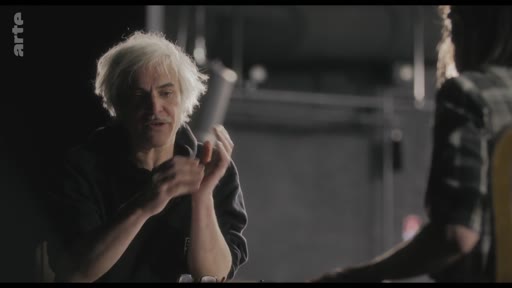 Un homme aux cheveux blancs, possiblement un metteur en scène ou un acteur, est filmé en plein échange lors d'une répétition artistique. L'ambiance est studieuse et concentrée, typique d'un travail de préparation théâtrale ou cinématographique.