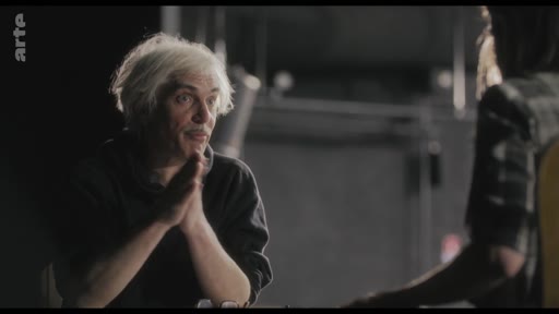 Un homme aux cheveux blancs, en plein échange lors d'une répétition artistique, s'exprime avec les mains. Il participe à une séance de travail ou de critique dans un cadre sombre.