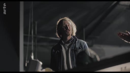 Un homme aux cheveux blonds, vêtu d'une veste en jean, s'exprime avec intensité lors d'une séance de répétition, illustrant une phase de tension dans le processus créatif.