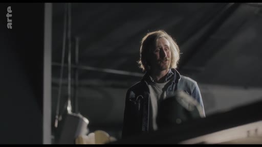Un homme aux cheveux blonds, visiblement en plein milieu d'une répétition, s'exprime dans un espace sombre. Cette scène illustre un moment de tension ou de pause lors d'un travail artistique.