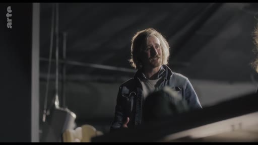 Un homme aux cheveux longs, vêtu d'une veste en jean, participe à une répétition dans un espace sombre, illustrant une phase de tension ou de travail créatif.