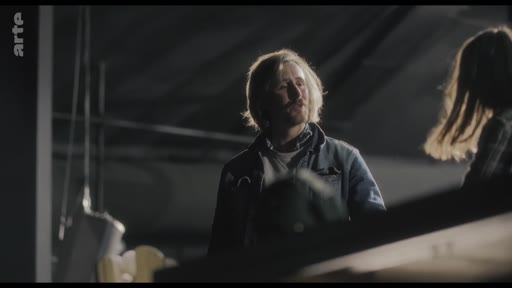 Un homme aux cheveux longs, possiblement un musicien ou un artiste, est filmé en plan moyen lors d'une répétition dans un espace sombre. L'ambiance suggère une tension ou une pause dans le travail créatif.