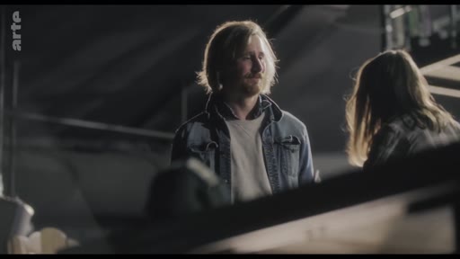 Un homme aux cheveux longs et à la barbe rousse, vêtu d'une veste en jean, est en pleine discussion avec une femme lors d'une répétition dans un espace sombre. L'ambiance semble tendue, illustrant une phase de travail artistique ou de préparation de spectacle.