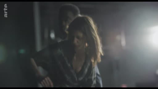 Une femme et un homme sont filmés dans une ambiance sombre et tamisée, évoquant une scène de fiction intense. L'éclairage dramatique souligne l'interaction entre les deux personnages dans ce qui semble être une séquence narrative.