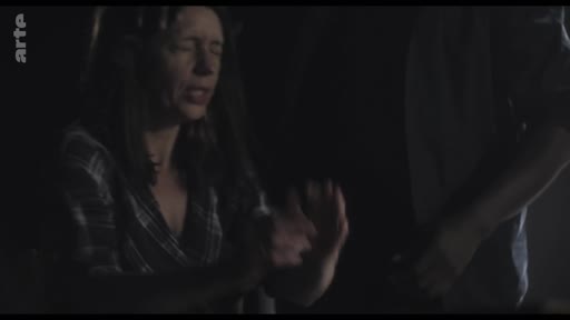 Une femme, filmée en gros plan dans une ambiance sombre, exprime une émotion forte ou une détresse lors d'une scène dramatique. L'éclairage met en valeur son visage et ses mains, soulignant l'intensité du moment.