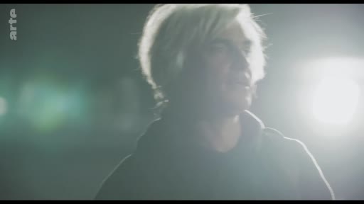 Un homme aux cheveux blonds, éclairé par une lumière vive en contre-jour, est filmé en gros plan dans une atmosphère sombre et dramatique, évoquant une scène de tension.