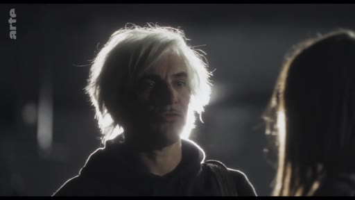 Un homme aux cheveux blonds, éclairé par un contre-jour, est filmé en gros plan lors d'une discussion tendue dans un environnement sombre.