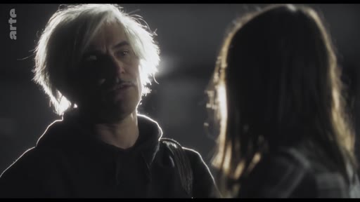 Un homme aux cheveux blonds ou gris, portant un sweat à capuche, est filmé en gros plan lors d'une conversation tendue dans une ambiance sombre et contrastée.