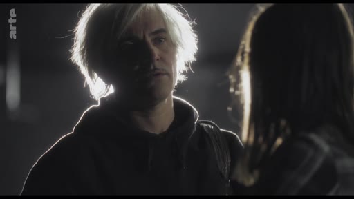 Un homme aux cheveux blonds, éclairé par un contre-jour, est en pleine conversation avec une autre personne dont on ne voit que le dos. La scène, filmée dans une pénombre marquée, évoque une interaction tendue ou intime.