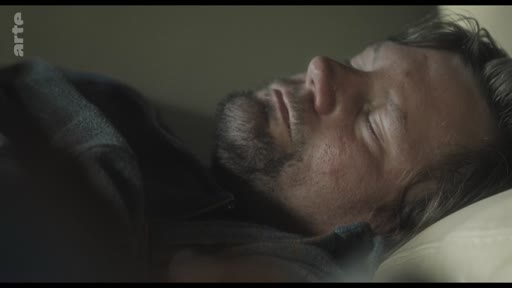 Un homme barbu est allongé et dort profondément dans un lit, dans une scène de repos ou de réflexion.