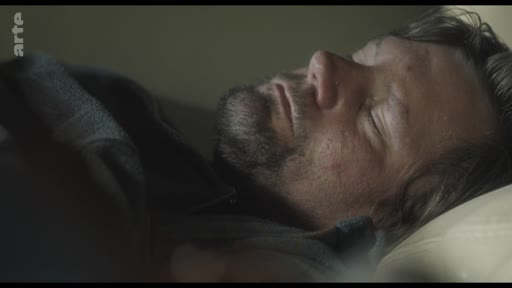 Un homme barbu est allongé et dort dans un lit, dans une scène de repos ou de réflexion.