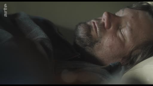 Un homme barbu est allongé et dort profondément dans un lit, dans une scène évoquant le repos ou la solitude.