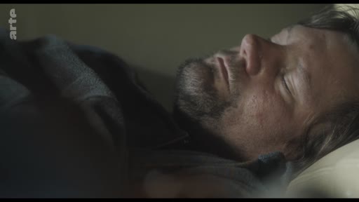 Un homme d'âge mûr avec une barbe courte est filmé en gros plan alors qu'il dort profondément dans un lit, dans une scène évoquant le repos ou la solitude.
