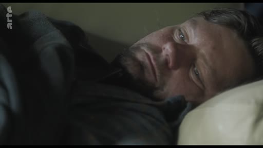 Un homme barbu est allongé sur un oreiller, le regard tourné vers le côté, dans une scène intime et mélancolique.