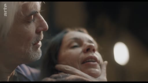 Un homme aux cheveux blancs tient fermement le visage d'une femme dans une scène de tension dramatique, illustrant une technique de jeu d'acteur.