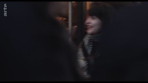 Une femme avec une frange et une écharpe à motifs sourit dans un espace urbain sombre, évoquant une réflexion personnelle ou une déambulation nocturne.