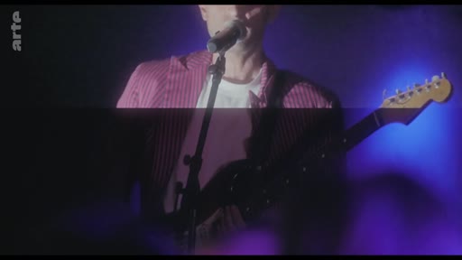 Un musicien vêtu d'une veste rayée rose chante devant un micro tout en jouant de la guitare électrique lors d'un concert. L'ambiance est marquée par un éclairage scénique bleu intense.