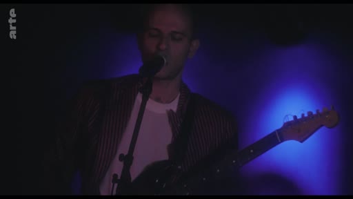 Un chanteur et guitariste se produit sur scène lors d'un concert, éclairé par des projecteurs bleus. L'artiste, vêtu d'une veste rayée, est capturé en pleine performance musicale dans une ambiance tamisée.