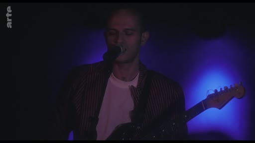 Un musicien chante et joue de la guitare électrique lors d'un concert filmé pour la chaîne Arte. L'artiste est éclairé par une lumière bleue intense dans une ambiance de performance live.