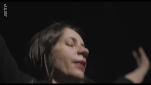 Une femme, filmée en gros plan sur un fond sombre, exprime une émotion forte avec les bras levés, dans une mise en scène dramatique.