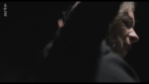 Une femme est filmée de profil dans une séquence de dramatisation télévisuelle, avec un éclairage sombre et dramatique.