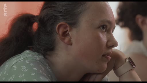 Une jeune femme, portant une montre connectée, écoute attentivement lors d'un atelier de réflexion. Elle est filmée en gros plan, exprimant une attitude concentrée et pensive.