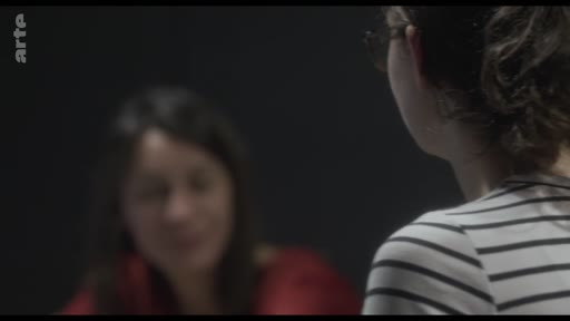 Une femme en t-shirt rayé est filmée de dos alors qu'elle discute avec une autre femme en arrière-plan, lors d'un entretien sur le théâtre et les émotions.