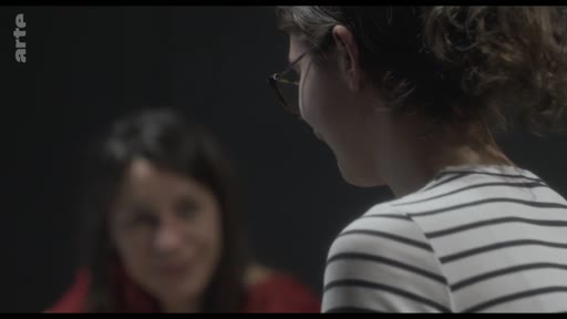 Une jeune femme aux cheveux bouclés et portant des lunettes est filmée de profil lors d'un entretien, avec une autre personne floue en arrière-plan. Cette séquence illustre une réflexion sur le théâtre et les émotions.