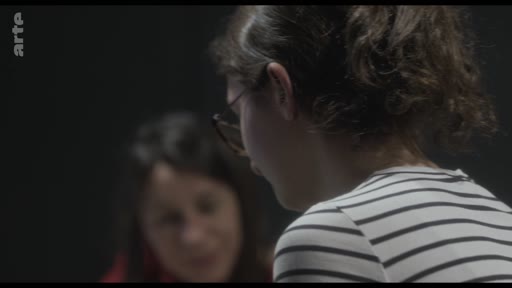 Une jeune femme aux cheveux attachés et portant des lunettes est filmée de profil lors d'une discussion, avec une autre personne floue en arrière-plan. Cette séquence illustre une réflexion sur le théâtre et les émotions.