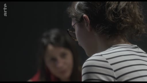 Une jeune femme aux cheveux attachés et portant des lunettes est filmée de profil, en discussion avec une autre personne lors d'un entretien sur le théâtre et les émotions.