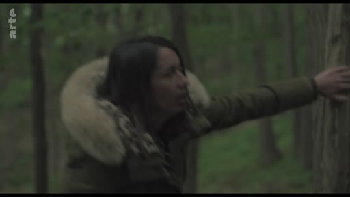 Une femme vêtue d'une veste à capuche en fourrure traverse une forêt dense et sombre, illustrant une scène de réflexion ou de recherche en extérieur.