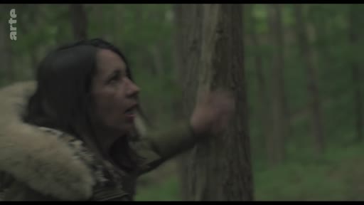 Une femme vêtue d'une veste à capuche en fourrure explore une forêt, touchant le tronc d'un arbre avec émotion.