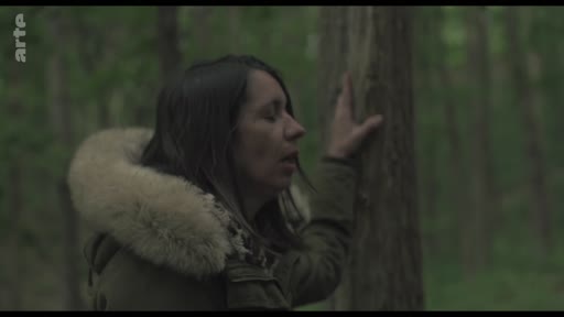 Une femme portant une veste à capuche en fourrure se tient dans une forêt, la main posée sur le tronc d'un arbre, dans une attitude de recueillement ou de réflexion.