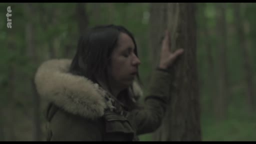 Une femme, vêtue d'une veste à capuche en fourrure, est filmée en plan moyen dans une forêt. Elle semble pensive ou émue, la main posée contre le tronc d'un arbre, dans une atmosphère mélancolique.