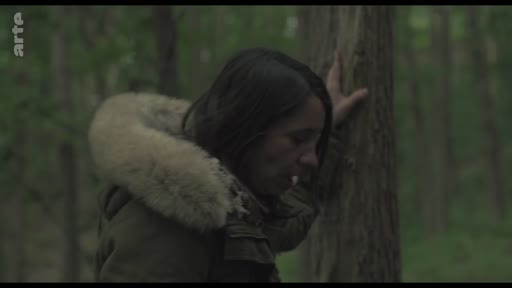 Une femme vêtue d'une veste à capuche en fourrure se tient dans une forêt, semblant pensive ou émotionnelle alors qu'elle s'appuie contre un arbre. Cette scène illustre un moment de réflexion solitaire en milieu naturel.