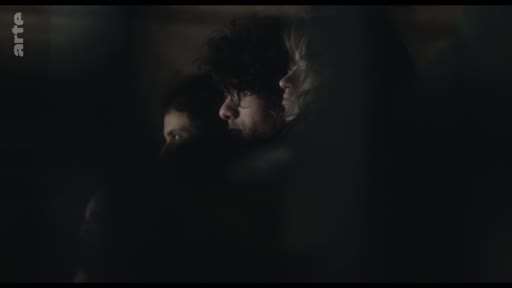 Trois individus, dont un homme aux cheveux bouclés, sont filmés dans une pénombre intense lors d'une séquence théâtrale ou cinématographique.