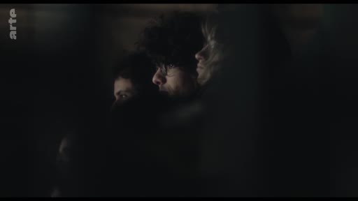 Trois individus, dont un homme aux cheveux bouclés, sont filmés en gros plan dans une pénombre quasi totale lors d'une performance théâtrale.