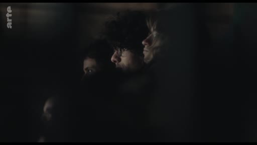 Un groupe de spectateurs, dont un homme aux cheveux bouclés, observe une représentation théâtrale dans une salle sombre. L'ambiance est feutrée et concentrée sur la scène.