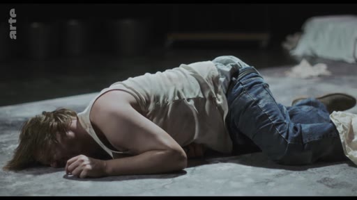Un homme en débardeur blanc et jean est allongé sur le sol d'une scène de théâtre, dans une mise en scène dramatique. L'image provient d'une captation de spectacle diffusée sur la chaîne Arte.