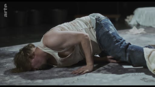 Un acteur, vêtu d'un débardeur et d'un jean, est allongé sur le sol dans une posture dramatique lors d'une représentation théâtrale capturée par Arte.